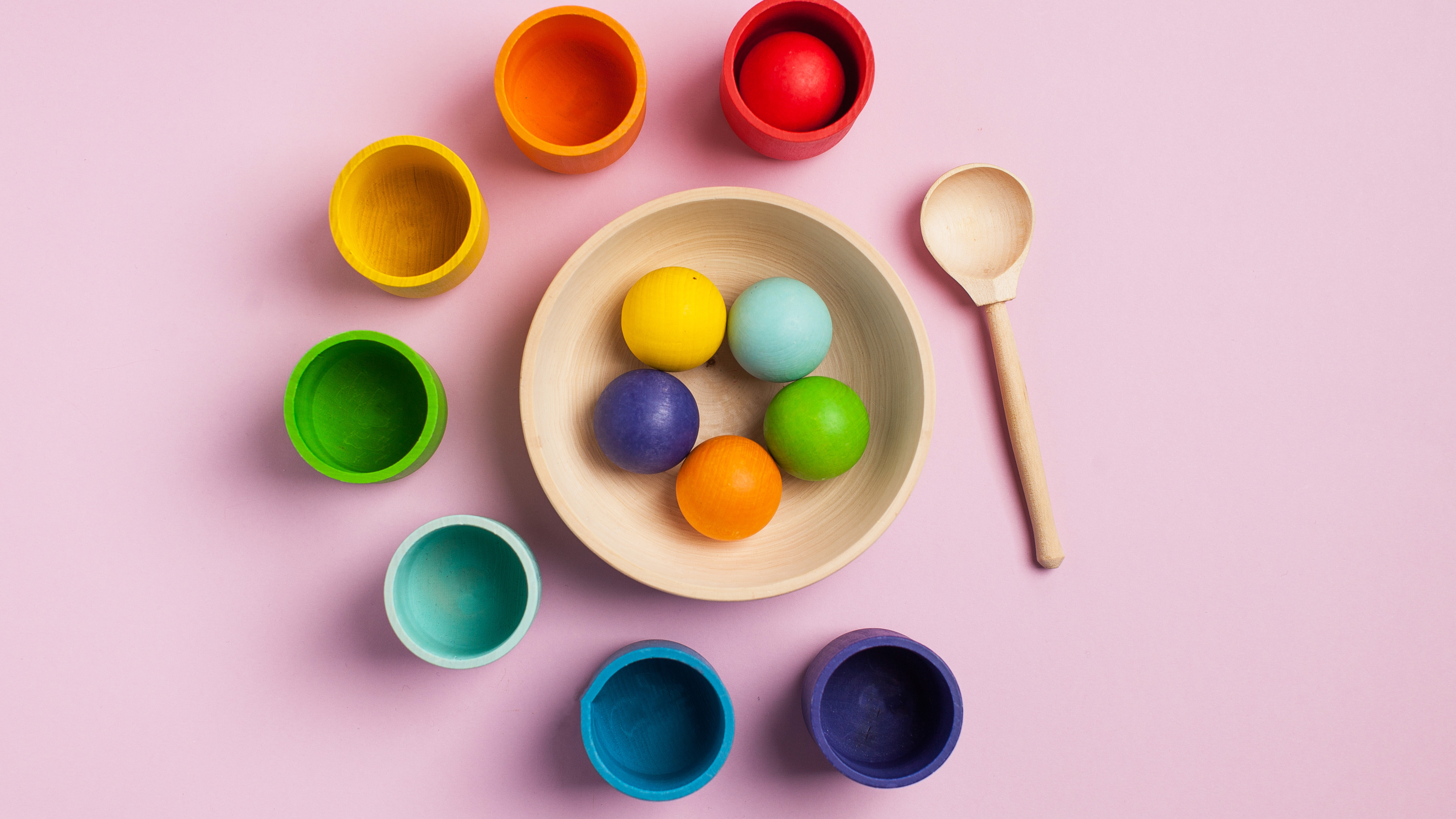 A flat lay of colorful wooden balls and matching cups on a pink background, illustrating a simple sorting game that is part of engaging and educational easy toddler activities at home.